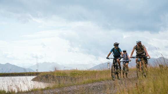 Three mountain bikers ride on a gravel path by a reservoir under cloudy skies near Bürglalmbahn in Dienten | © Hochkönig Bergbahnen