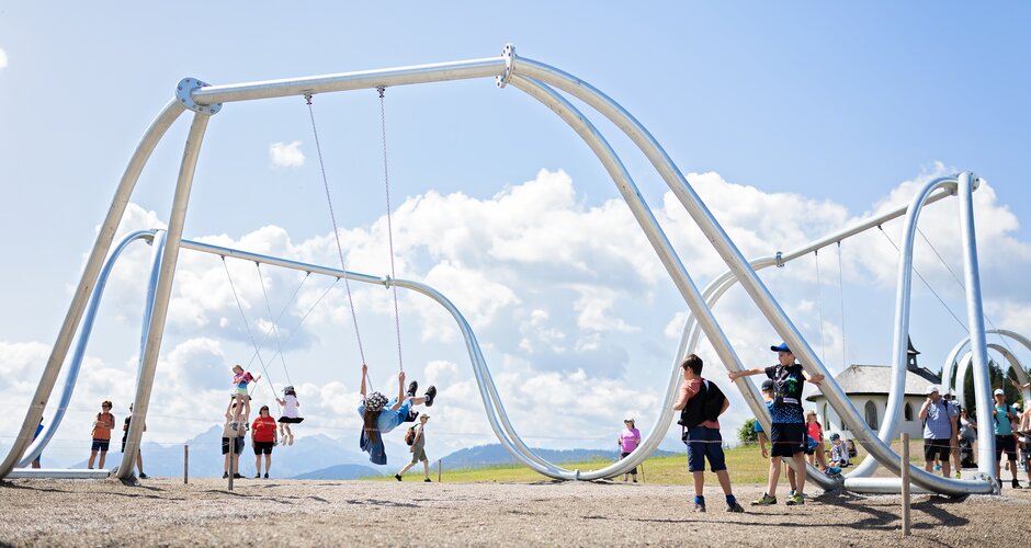 Kids swinging on oversized swings at Hochkönig swing park with Alpine backdrop on a sunny day | © Manu Lochner