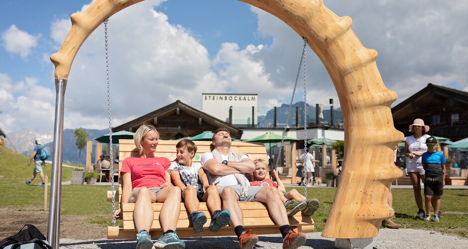Family relaxing on large wooden swing with carved arch at Steinbockalm in Hochkönig swing park | © Manu Lochner