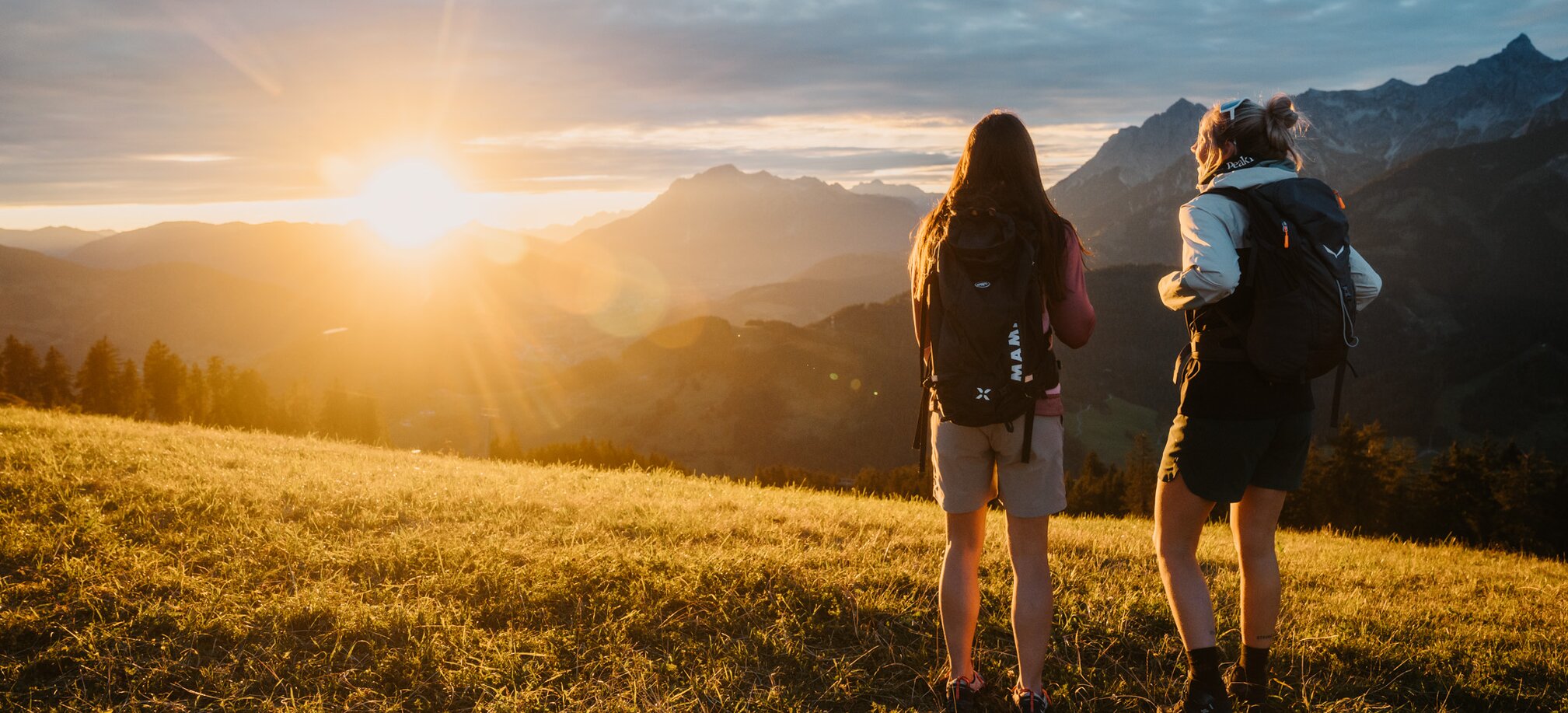 Two female hikers stand on a grassy slope in Dienten looking at mountains near Gabühelbahn at sunset. | © Hochkönig Bergbahnen