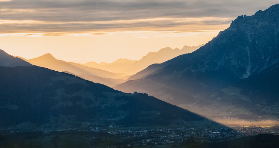 View over Dienten across a valley with mountain silhouettes, sun rays and a cloud-filled sky in morning light. | © Hochkönig Bergbahnen