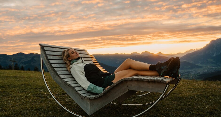 A woman lies on a wooden lounge bench on a grassy slope in Dienten with mountain views at sunset. | © Hochkönig Bergbahnen
