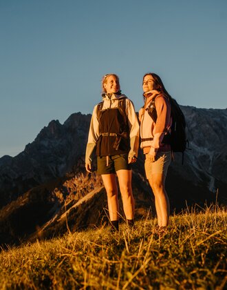 Two female hikers stand on a grassy slope in evening light in Dienten with a rocky mountain massif near Gabühelbahn. | © Hochkönig Bergbahnen