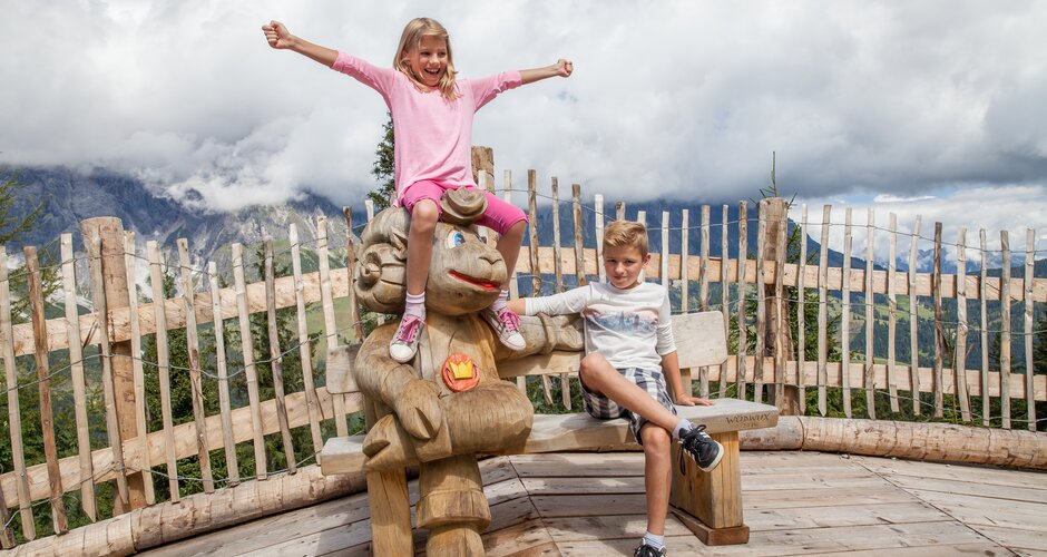 Two kids laugh and pose on carved wooden mascot bench at alpine play park with scenic mountain view behind