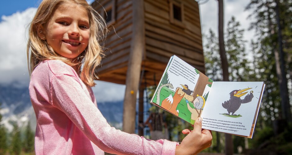 Girl shows an open children’s book in front of a wooden hut at Hochkönig play park with trees and mountains behind