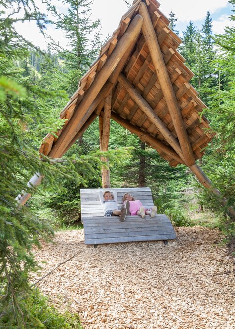 Two kids relax on a wooden lounger with roof in the shady forest of Hochkönig’s alpine play park