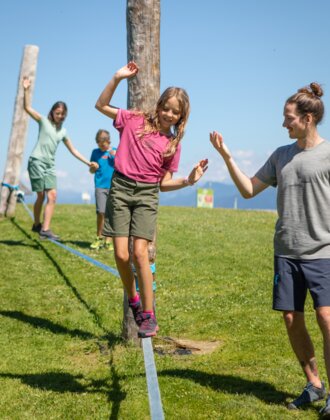 A girl balances on a slackline in Mühlbach while a man helps and two children follow behind. | © Hochkönig Bergbahnen, Roland Haschka