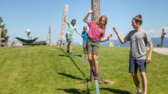 A girl balances on a slackline in Mühlbach while a man helps and two children follow behind. | © Hochkönig Bergbahnen, Roland Haschka