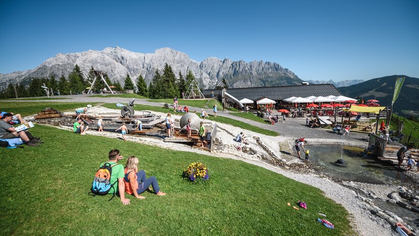 Spacious alpine playground in Mühlbach with water area, terrace, visitors and mountain massif behind. | © Hochkönig Bergbahnen