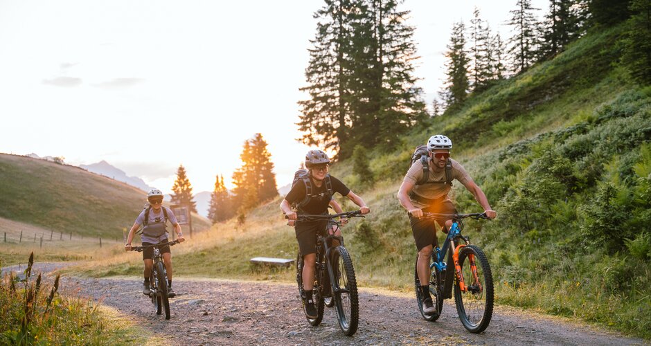 Three mountain bikers ride on a gravel path in Mühlbach at sunset near Karbachalmbahn and tall trees. | © Hochkönig Bergbahnen