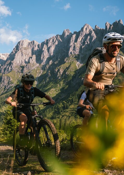 Three mountain bikers ride in Mühlbach on a trail in front of a steep rocky massif near Karbachalmbahn. | © Hochkönig Bergbahnen