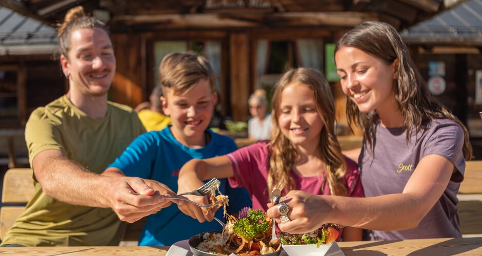 Four people sit at a wooden table outside an alpine hut and share a dish with salad in the sun. | © Hochkönig Bergbahnen, Roland Haschka