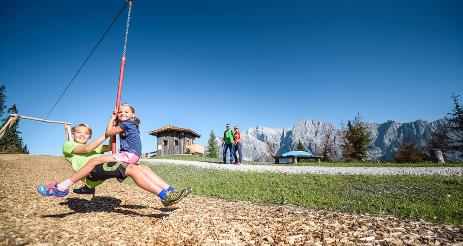 Two kids laughing on zipline at alpine playground, with the Hochkönig mountain range in the sunny background