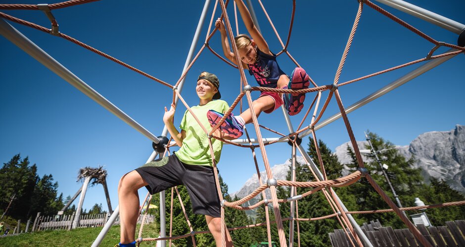 Two kids climbing rope net at playground under blue sky with alpine mountain backdrop in Hochkönig