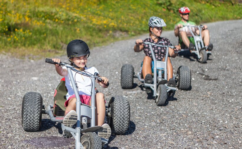 Child and adults riding mountain carts with helmets on a gravel road in sunny alpine landscape
