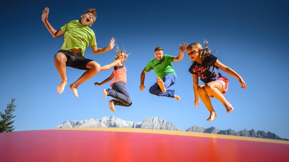 Four people joyfully jumping on an air cushion with alpine backdrop and blue sky at Hochkönig