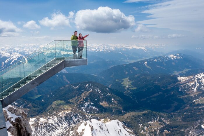 Two people stand on the glass Skywalk at Dachstein, overlooking snow-covered peaks and a vast mountain landscape. | © Harald Steiner