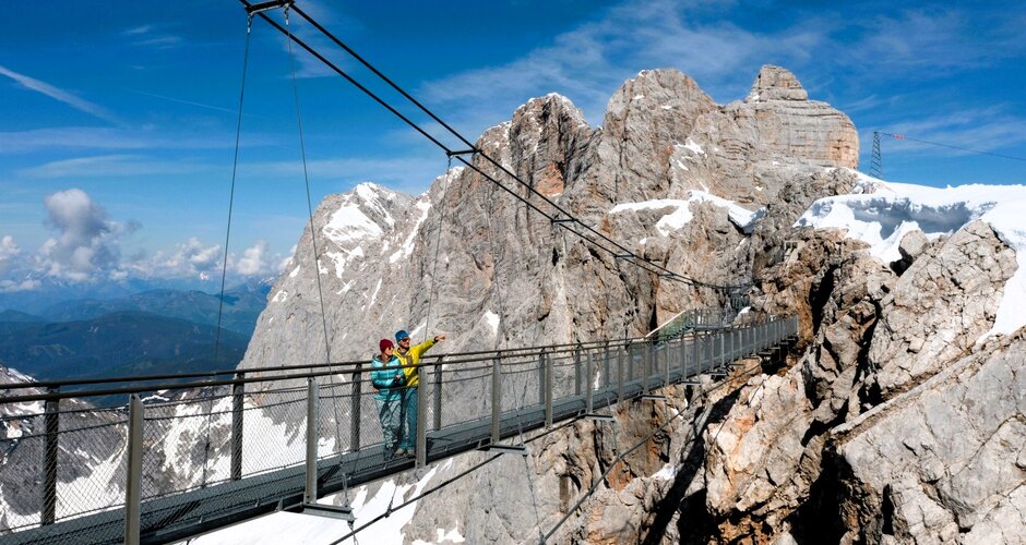 Two people cross the Dachstein suspension bridge with snowy peaks and rugged cliffs in the background under clear blue skies. | © Harald Steiner