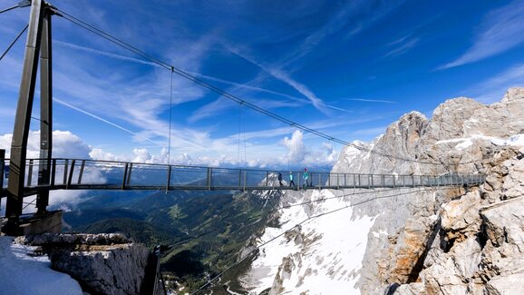 People cross a suspension bridge on snowy Dachstein with a wide view over green valley under a clear blue sky. | © Harald Steiner