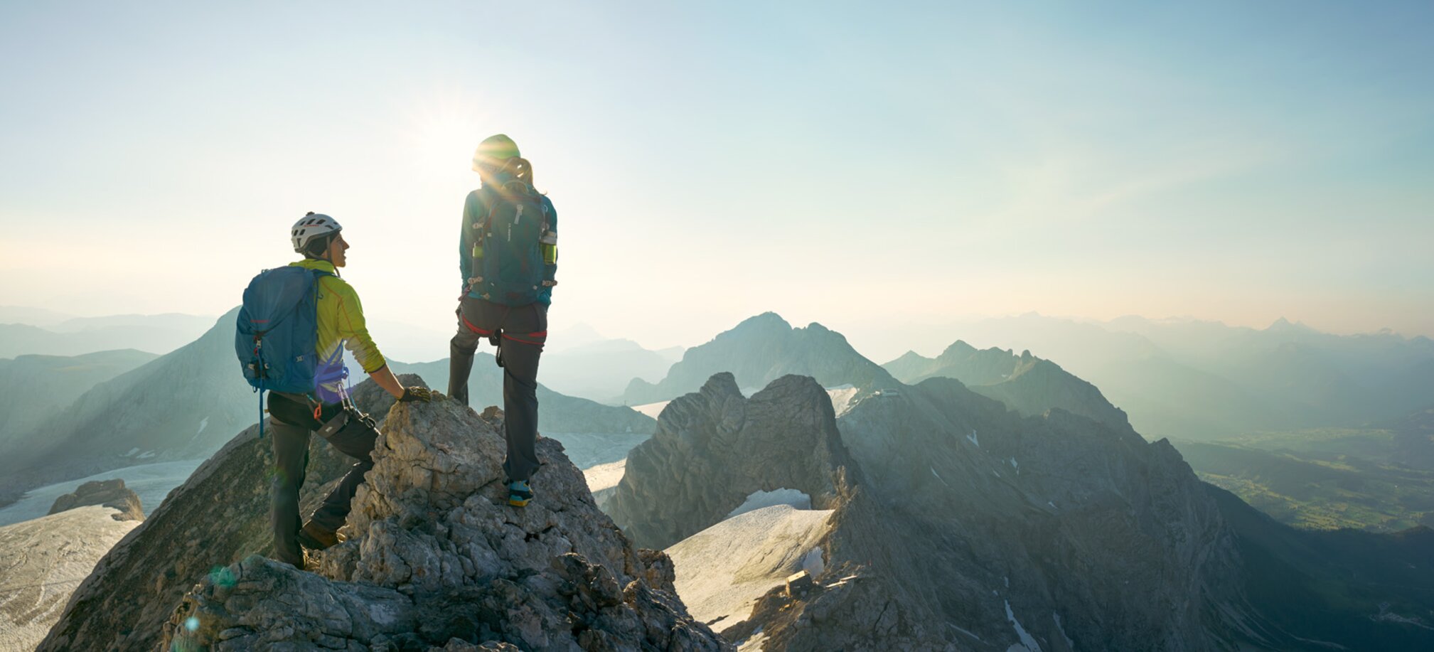Two climbers with backpacks and helmets stand on Dachstein summit, sunrise and alpine panorama unfolding in the background. | © Peter Burgstaller