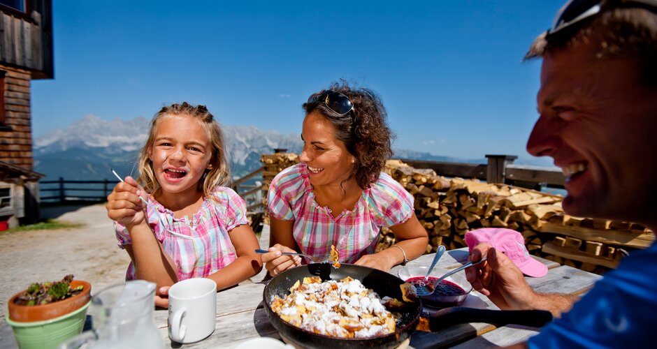 Family eating Kaiserschmarrn on a sunny terrace with mountain hut flair and view of the Hochwurzen range. | © Tom Lamm
