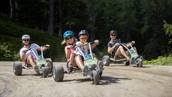 Three adults and one child laugh while riding mountain go-karts on a forest trail in sunny weather. | © Harald Steiner