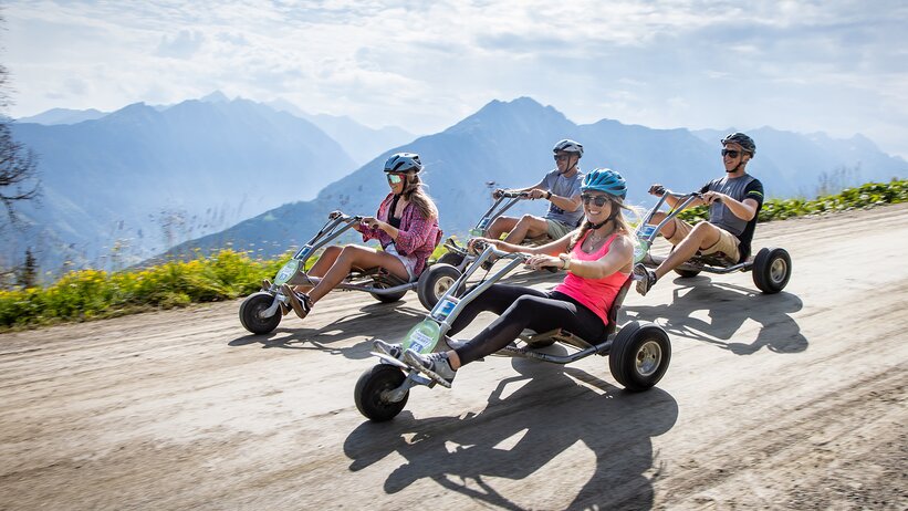 Four people ride mountain go-karts downhill on gravel road with alpine views and a summer vibe. | © Harald Steiner