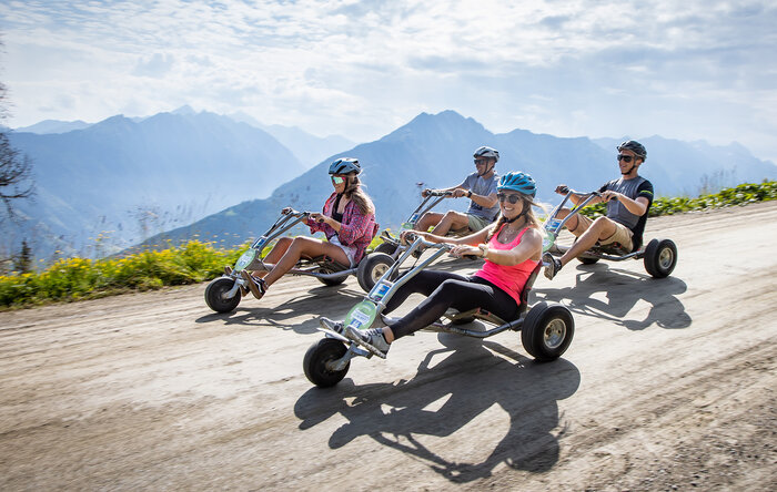 Four people ride mountain go-karts downhill on gravel road with alpine views and a summer vibe. | © Harald Steiner
