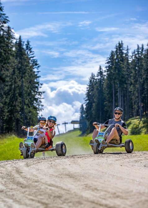 Two adults and a child ride mountain go-karts down a gravel road between forest and alpine meadow. | © Harald Steiner