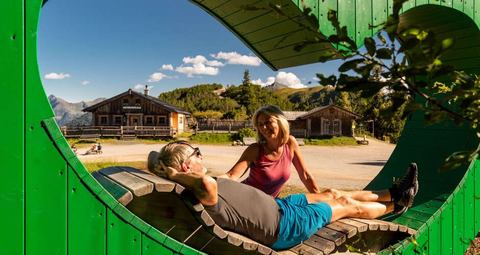 Hiking couple relaxes on heart-shaped bench with view of alpine huts and sunny mountain scenery. | © Christine Höflehner