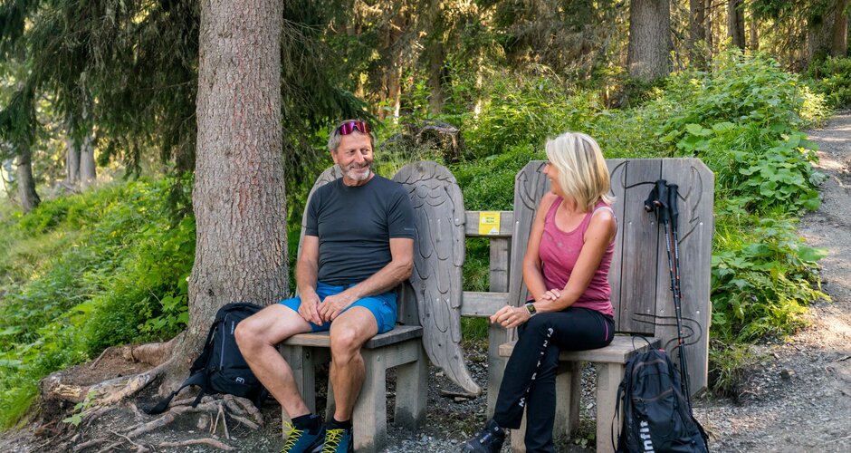 Hiking couple takes a break on carved wooden benches with wing motifs along the shady Bankerlweg trail. | © Christine Höflehner