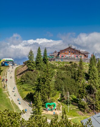 Go-kart riders and hikers at Hochwurzen summit with alpine hut, heart-shaped spot and mountain view. | © Planai/Kovacsics