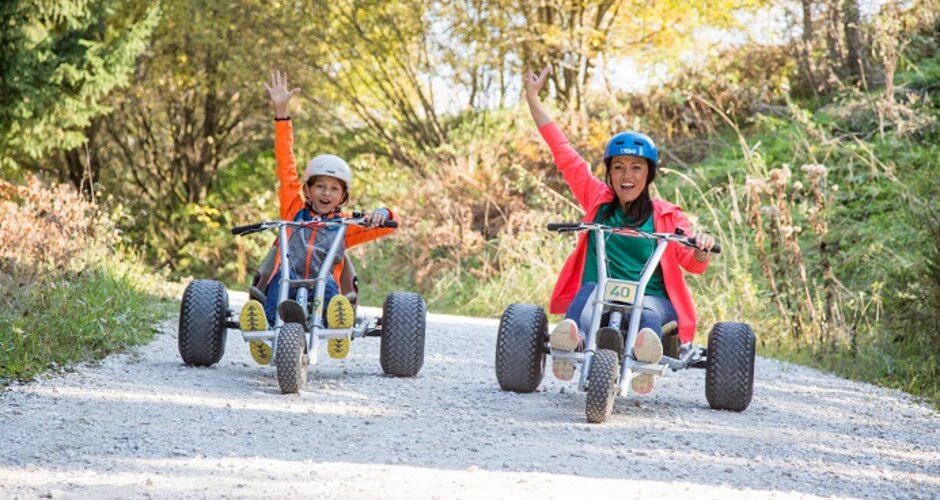 Woman and child ride mountain karts on gravel path, smiling and raising arms in excitement. | © Elisabeth Wolkenstein
