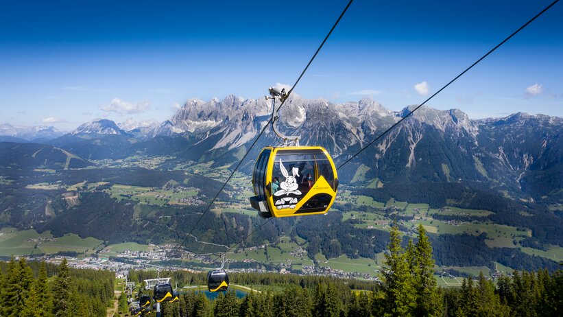 Yellow gondola with Hopsiland bunny above Planai with Dachstein mountains and green Enns valley in bright summer weather. | © Josh Absenger