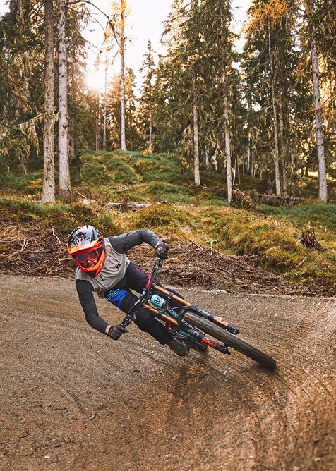A mountain biker in helmet and goggles carves a sharp bermed turn on a forest trail in the bike park during golden sunlight. | © Armin Walcher