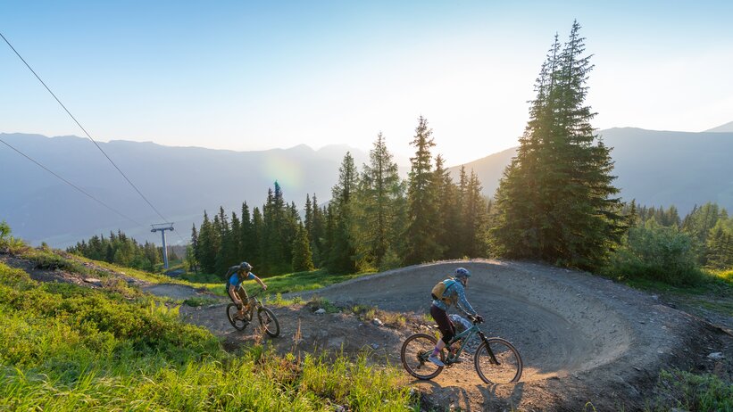 Two mountain bikers ride a bermed curve at sunset on a forest trail with alpine scenery and lush mountain landscape. | © Christoph Oberschneider