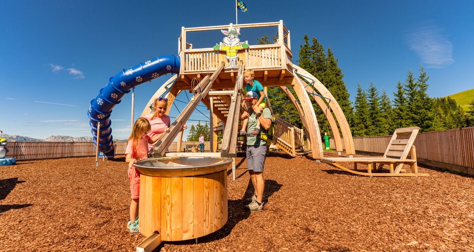 Family plays at Hopsiland Planai with water chute in front, wooden play tower with blue slide and bunny figure in background. | © Christine Höflehner