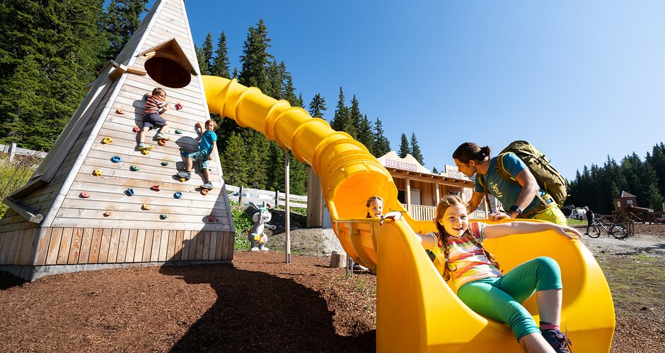 Kids climb a wooden play tower with yellow slide at Hopsiland Planai, one girl slides down smiling, father standing next to her. | © Josh Absenger