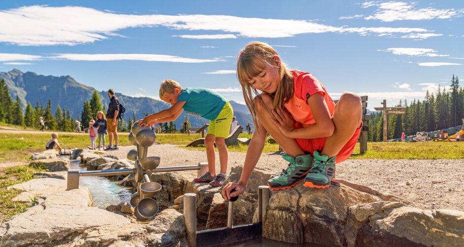 Two kids play at a water game station with channels and gates at Hopsiland Planai, mountains and trees in the background. | © Christine Höflehner