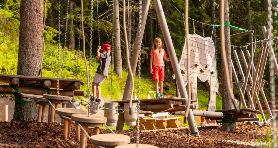 Two kids balance on wooden climbing elements at Hopsiland Planai, surrounded by forest and playful animal-shaped structures. | © Christine Höflehner