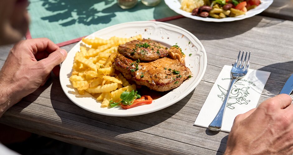 Grilled meat with fries, salad and drinks on wooden table in sunlight, served on a summer hut terrace on Planai mountain. | © Dietmar Körbler