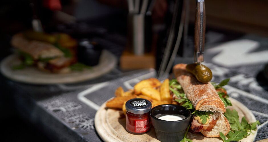 Crispy baguette with bacon, rocket, sauces, pickle and wedges served on a wooden plate in bright sunlight on a black tablecloth. | © Dietmar Körbler