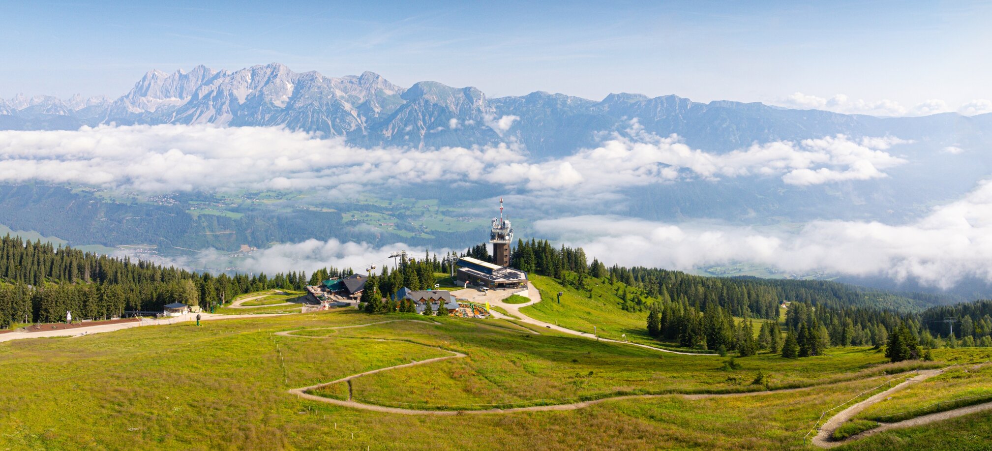 Vast alpine pasture with a mountain station and observation tower, surrounded by meadows, forest, and mountain ranges. | © Josh Absenger