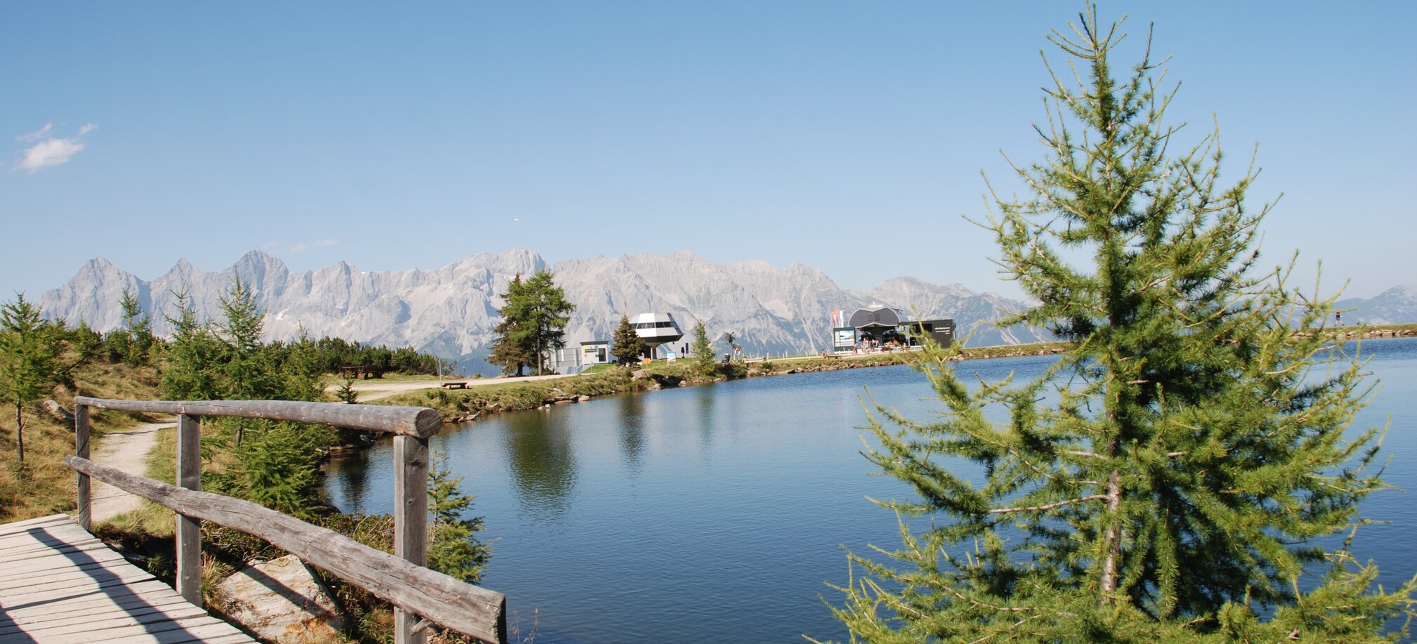 Ein Steg führt am ruhigen Bergsee der Reiteralm entlang, im Hintergrund das Dachsteinmassiv und zwei Bergstationen bei Sonne. | © Reiteralm Bergbahnen