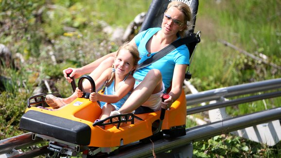 Mother and daughter ride the Rittisberg alpine coaster together, smiling while racing downhill in a summer toboggan car. | © photo-austria