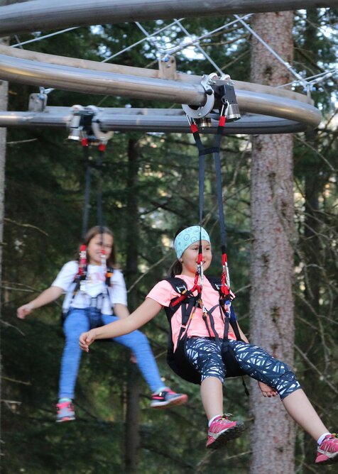 Two children glide safely in harnesses on the Flyline ride through the forest at Rittisberg, surrounded by tall pine trees. | © Erlebnis Rittisberg