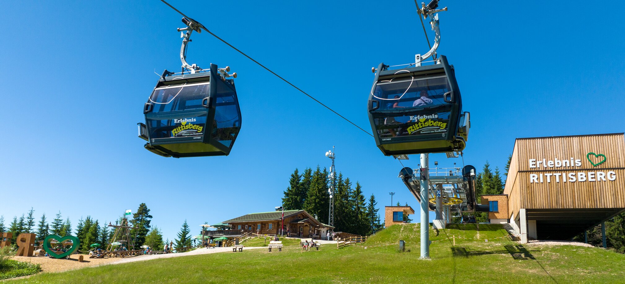 Two gondolas of the Rittisberg cable car reach the top station, with play area, huts and blue summer sky in the background. | © Rittisberg