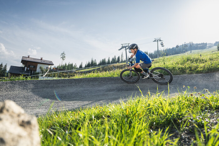 A boy in helmet rides a Woom bike on a curved trail with an alpine house, grassy slope, and gondola lift in the background. | © Snow Space Salzburg - Chris Eder