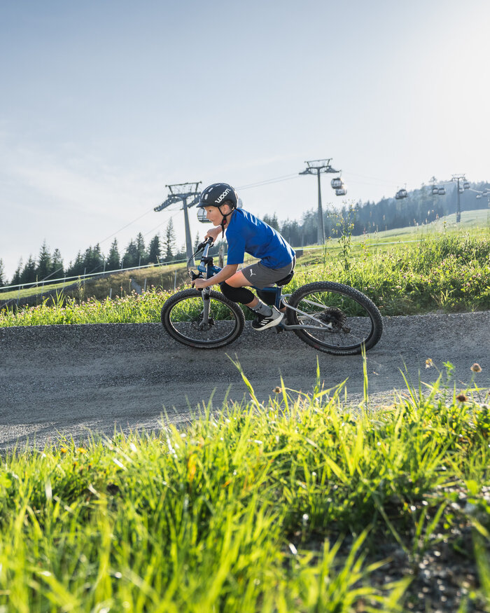 Ein Junge mit Helm fährt ein Woom-Bike auf einem Schottertrail, im Hintergrund sind grüne Wiesen, Wald und Gondeln sichtbar. | © Snow Space Salzburg - Chris Eder