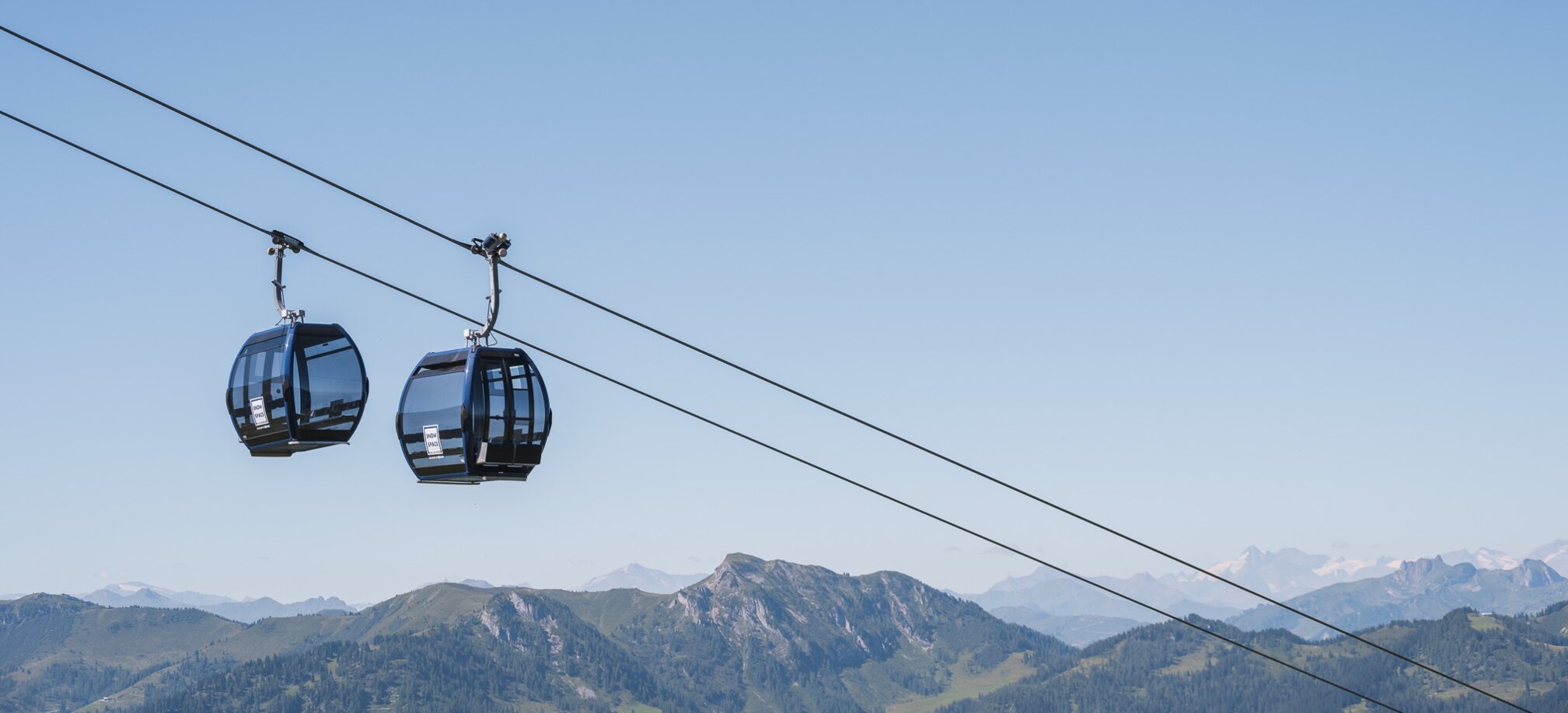 Two black panoramic gondolas float above green summer mountains, with snowy Alpine peaks in the far background. | © Snow Space Salzburg